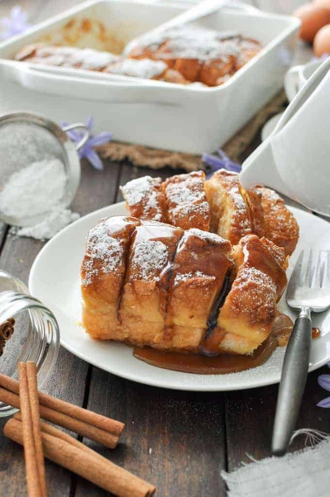Hasselback Baked French Toast on a white plate, dusted with icing sugar and drizzled with maple syrup.