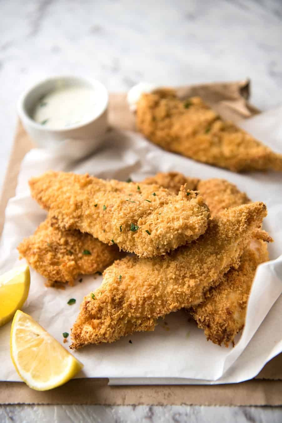 A pile of Oven Fried Chicken Tenders with lemon wedges and ranch dipping sauce in the background.