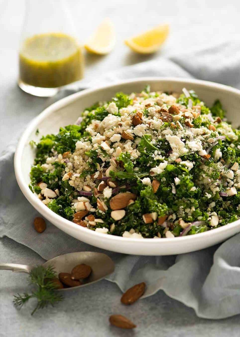Photo of Kale and Quinoa Salad in a white rustic salad bowl with a small jug of lemon dressing on the side.
